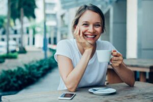Happy Nice Lady Drinking Coffee in Outdoor Cafe Closeup portrait of smiling young beautiful woman looking at camera, leaning head on hand and drinking coffee at cafe table outdoors with street view in background. Front view.