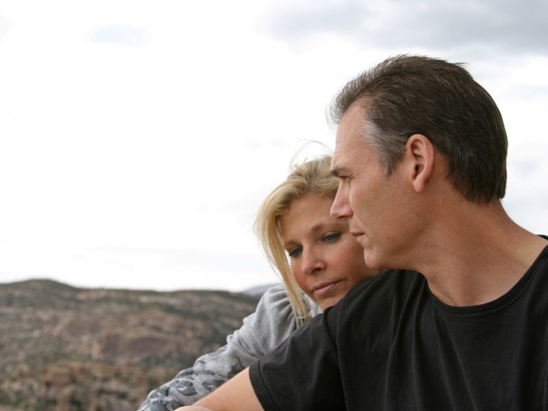 "A older adult married couple takes a break while hiking in the mountains. They are enjoying the view and enjoying each other's company. Slightly smiling and looking off into the distance, however they look a bit solemn and serious."