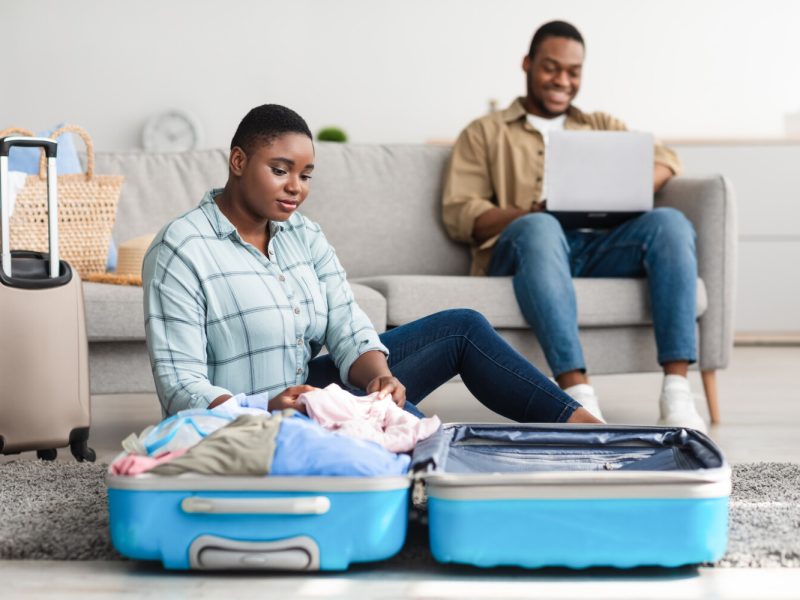 Vacation Preparation. Black Woman Packing Suitcase While Husband Booking Tickets On Laptop Computer Sitting At Home. Family Preparing For Travel Abroad. Selective Focus