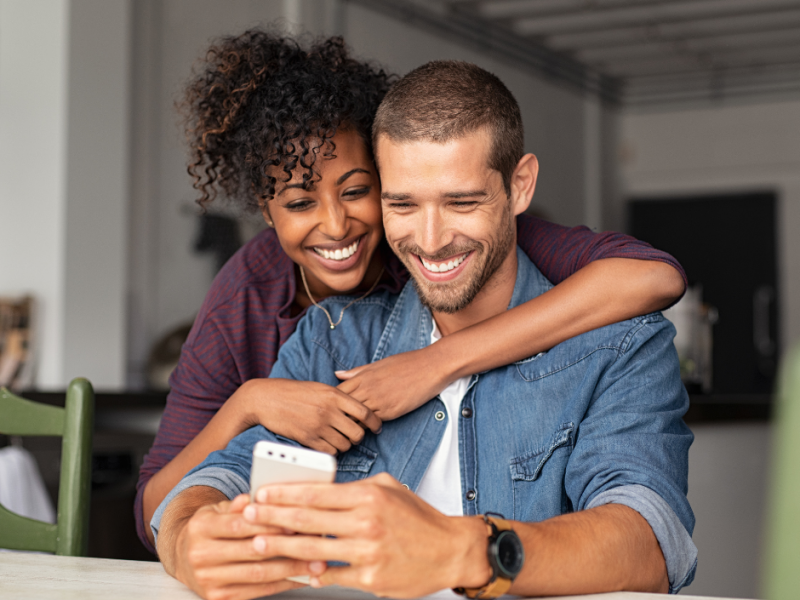 Happy bi racial couple looking at mobile phone after apply for patient fi financing for MCRM Fertility